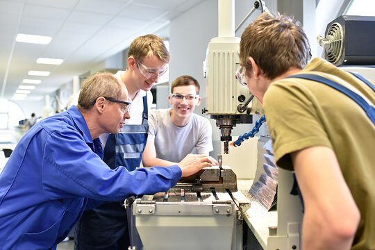 Ein Meister erklärt in der Werkstatt drei Auszubildenden einen Arbeitsschritt.