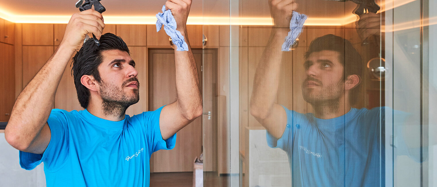 Mann im blauen T-Shirt benutzt eine Kartuschenpistole, um Dichtmasse an einer Glaswand aufzutragen; seine Arbeit spiegelt sich im Glas wider.